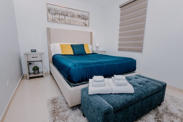 Un dormitorio con cama, mesita de noche, lámpara y un cuadro en la pared.