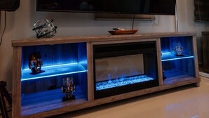 Una sala de estar con un televisor de pantalla plana grande montado en la pared, un mueble de madera para entretenimiento con estantes, y una vitrina de vidrio con objetos decorativos.