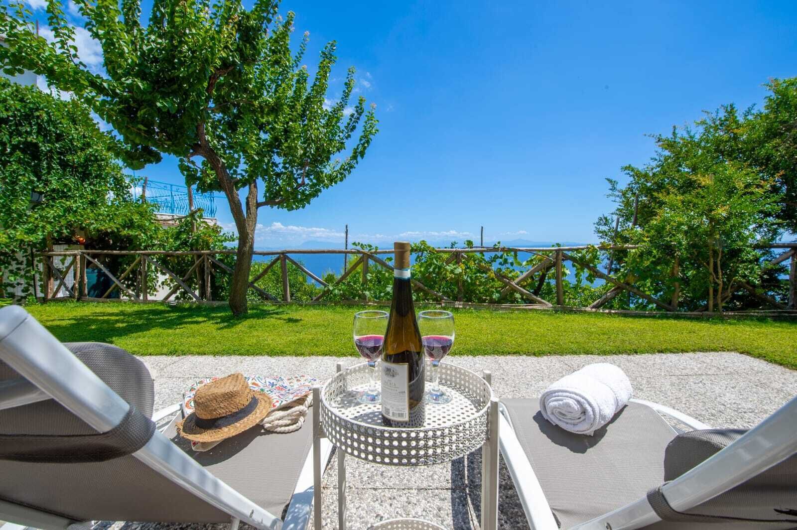 Ein Tisch mit einer Weinflasche, Gläsern, einem Strohhut und einem Handtuch auf einer Terrasse mit Blick auf eine Wiese und ein Gewässer.