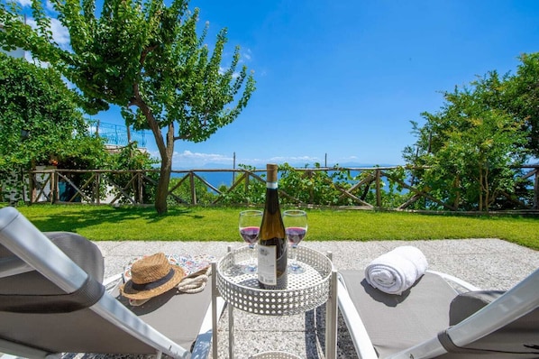 A table with a bottle of wine, glasses, a straw hat, and a towel on a deck overlooking a grassy area and a body of water.