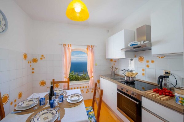 Une cuisine avec vue sur la mer, une table à manger dressée avec des assiettes et des verres, et un fourneau.
