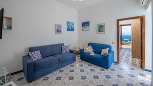 Ein Wohnzimmer mit zwei blauen Sofas, einem Musterfliesenbelag und Blick auf das Meer durch eine offene Tür.