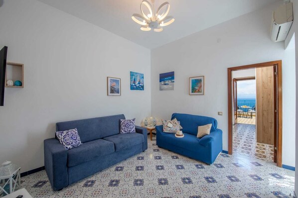 A living room with two blue sofas, patterned floor tiles, and a view of the sea through an open door.