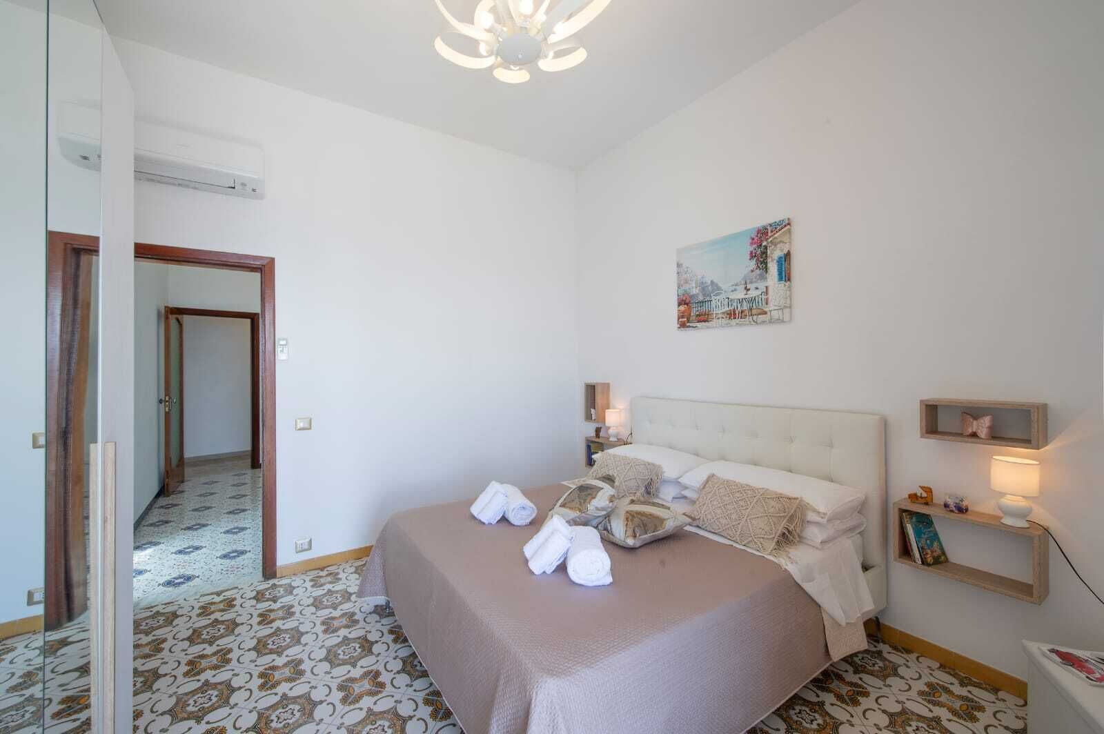 A bedroom with a bed, bedside lamps, a painting, and a small shelf.