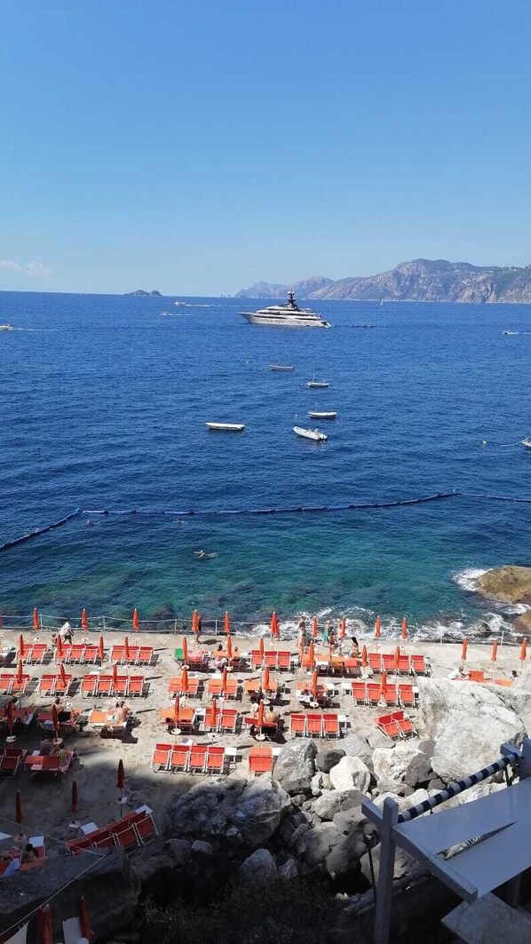 Una playa con hileras de sillas y sombrillas anaranjadas, terreno rocoso y barcos en el agua.