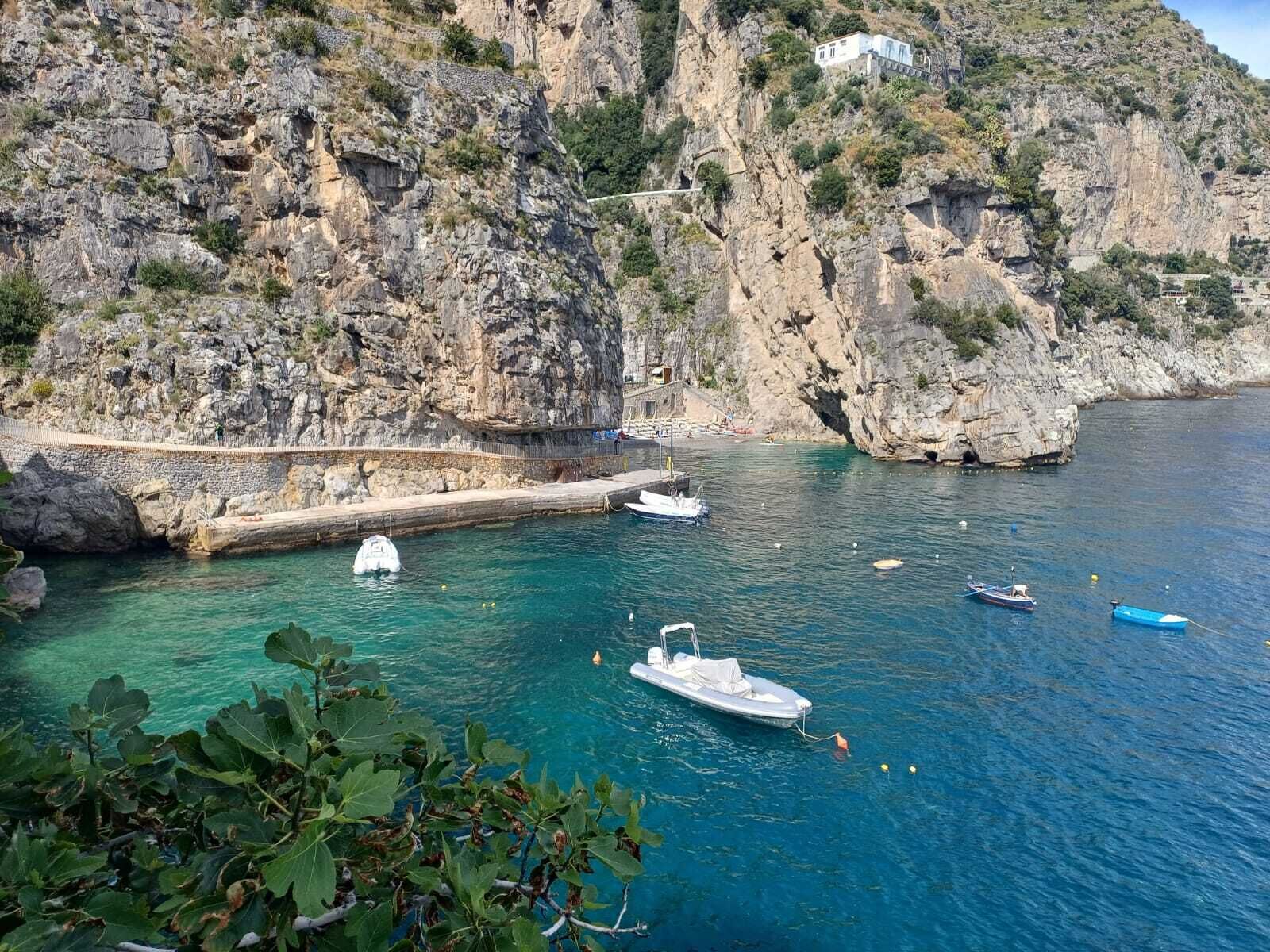 A coastal scene with rocky cliffs, a clear blue sea, and boats anchored near the shore.