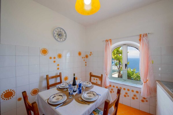 A dining area with a table set for four, a clock on the wall, and a window with a view of the sea.
