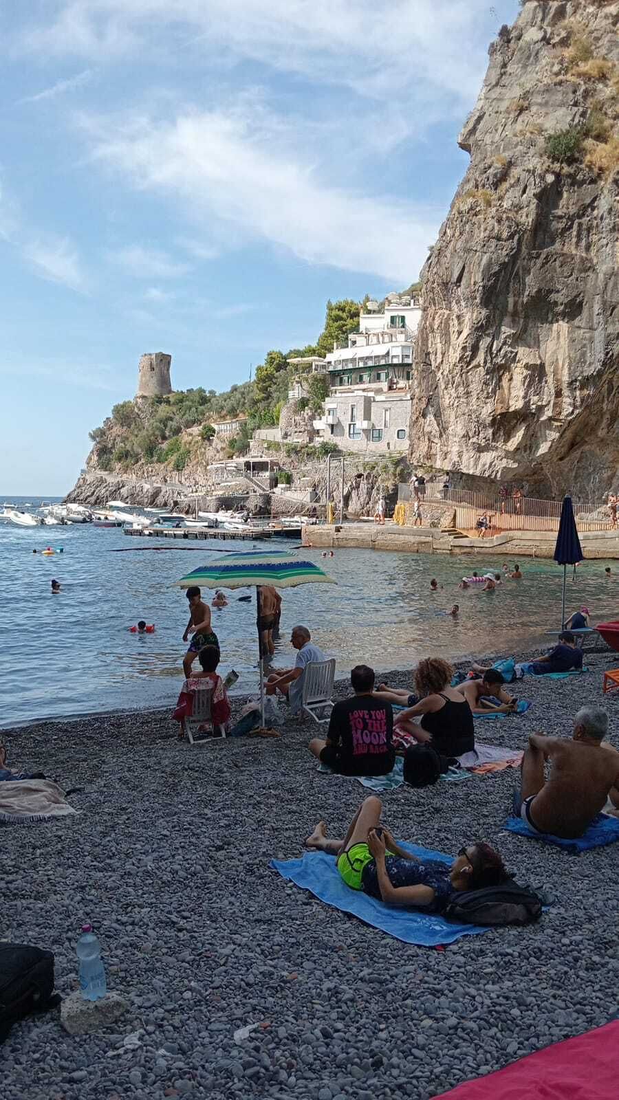 Une plage où des gens se prélassent au soleil et se baignent, un château perché sur une falaise et des bateaux sur l’eau.
