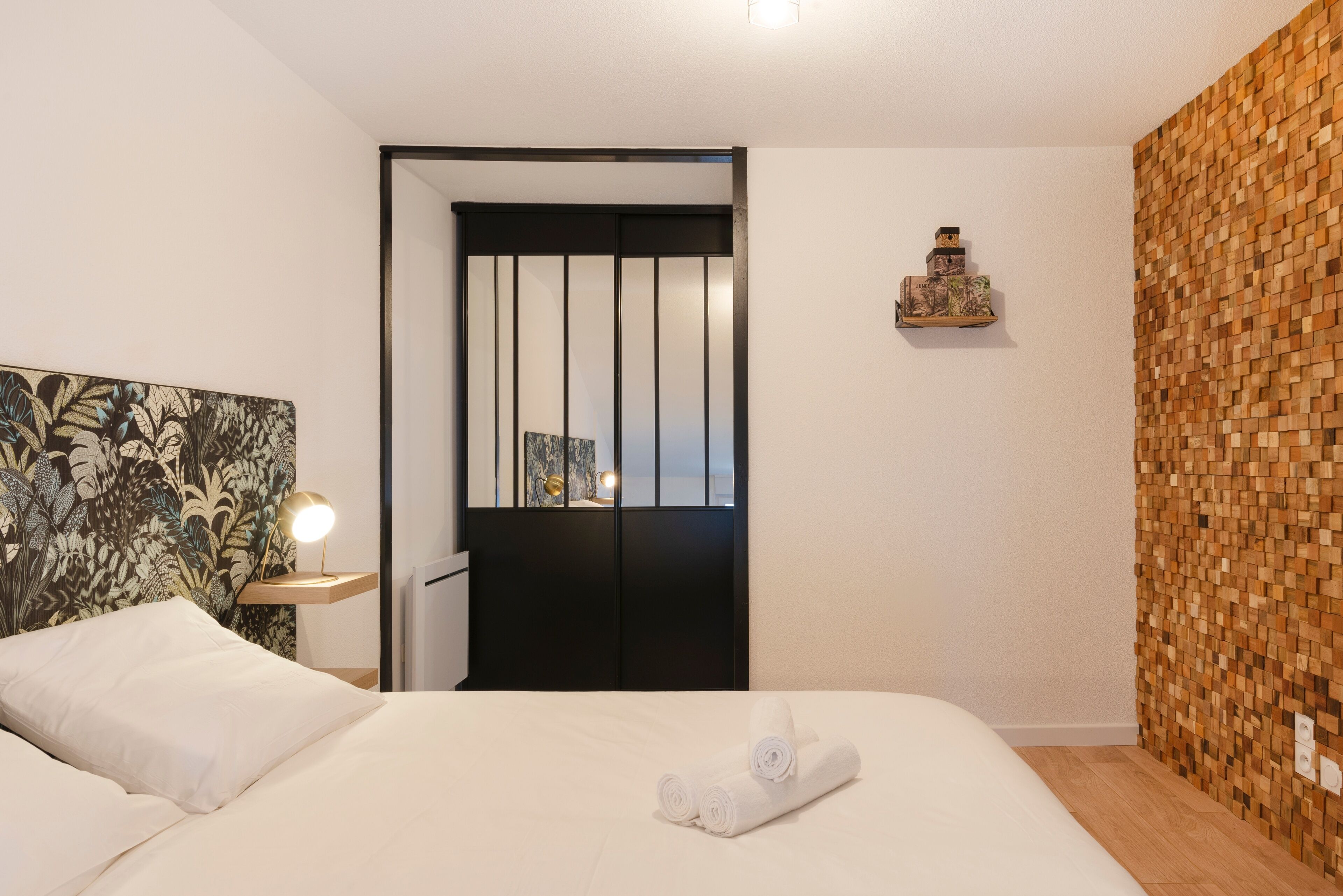 A neatly made bed with white linens and a rolled towel, a bedside table with a lamp, a wall with a wooden pattern, and a door with vertical blinds.
