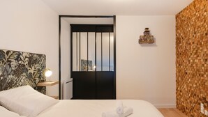 Un lit bien fait, avec du linge de lit blanc et une serviette enroulée, une table de chevet avec une lampe, un mur orné d’un motif en bois et une porte avec des stores verticaux.
