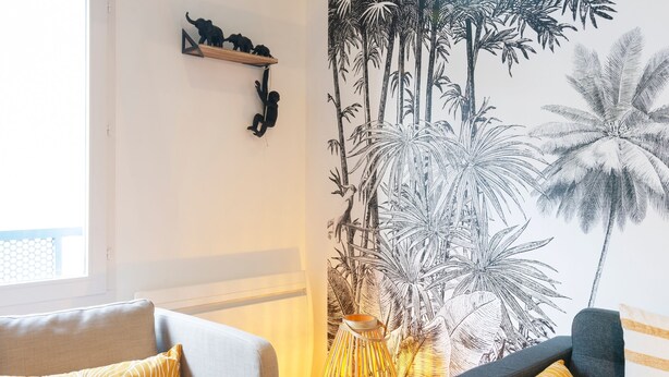 Un salon avec deux canapés, une fresque murale représentant une végétation tropicale, un plafond en bois et une petite étagère ornée de figurines.