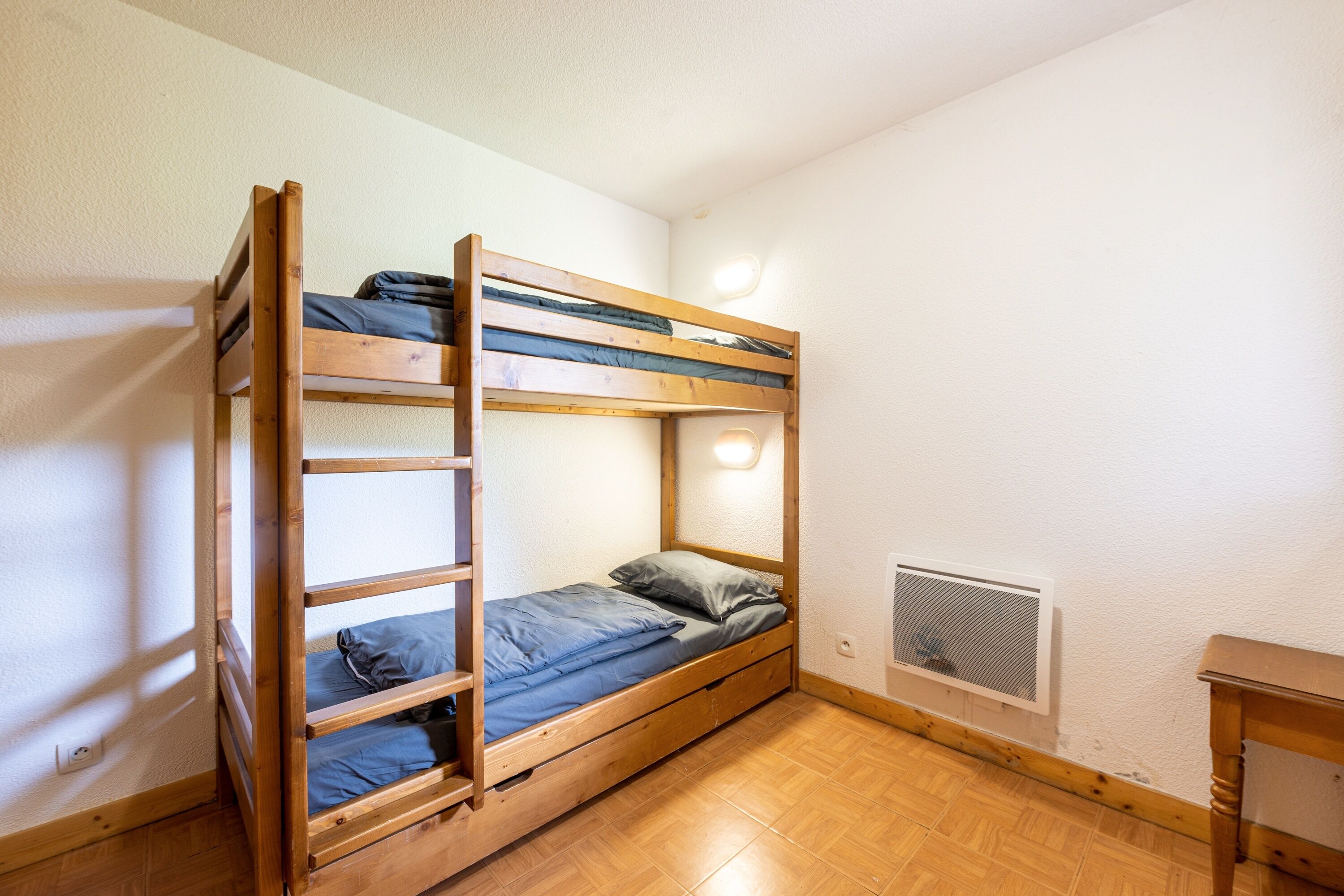 Una cama litera con colchón, almohadas y cabecera. Hay una mesita y un calentador fijado a la pared.