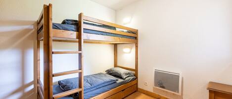Una cama litera con colchón, almohadas y cabecera. Hay una mesita y un calentador fijado a la pared.
