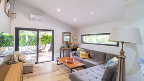 A modern living room with a grey sofa, wooden coffee table, and a lamp. There is a sliding glass door leading to an outdoor area with plants and furniture.