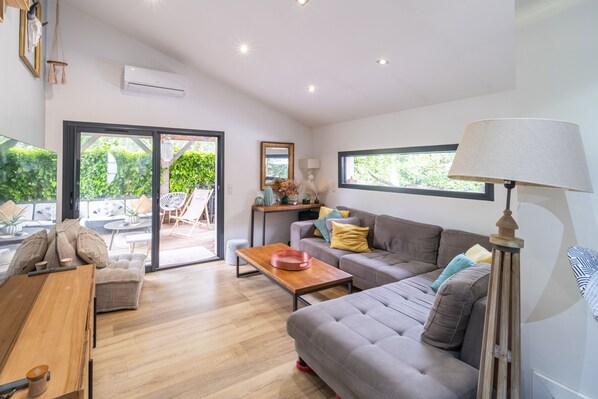 A modern living room with a grey sofa, wooden coffee table, and a lamp. There is a sliding glass door leading to an outdoor area with plants and furniture. - La Maison Riquet - Maison avec jacuzzi (Labège)