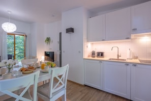 A modern kitchen with white cabinets, a dining table with chairs, a hanging light fixture, and a window with curtains.