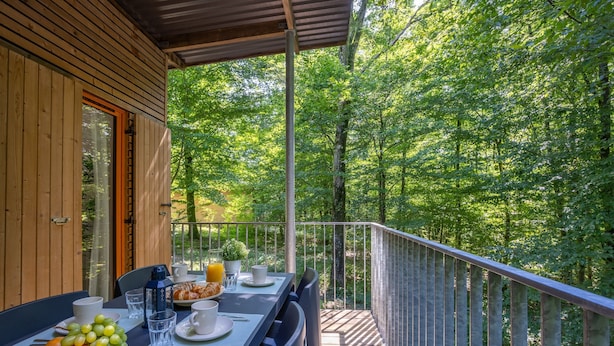 Una terraza de madera con una mesa dispuesta para el desayuno, rodeada por una barandilla metálica, con vistas a un bosque frondoso.