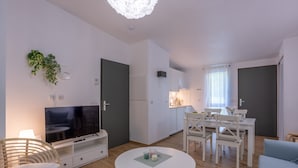 Una sala de estar moderna con televisor, muebles blancos, un comedor y una cocina.