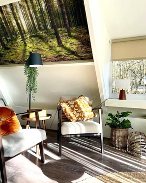Una sala moderna con un gran mural de un bosque, una planta colgante, un sillón cómodo con un cojín estampado y vistas a árboles a través de una ventana.