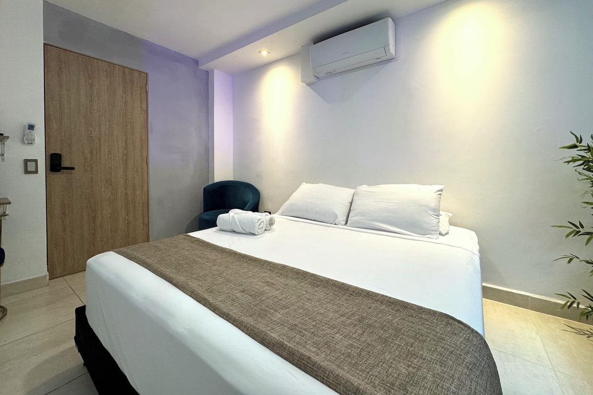 Une chambre d’hôtel comprenant un lit, une chaise, un appareil de climatisation, une porte et un luminaire fixé au mur.