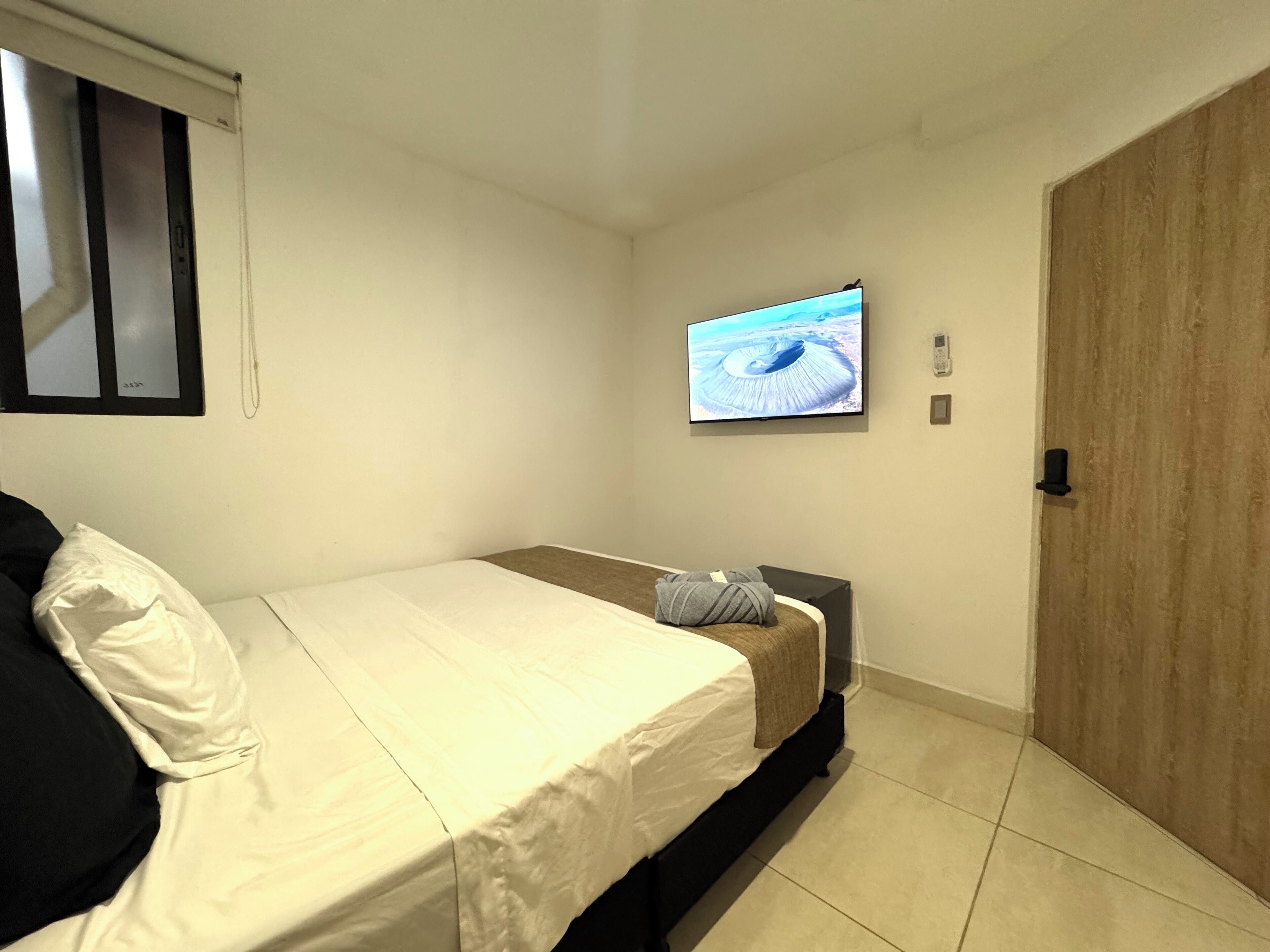 A small, clean room with a bed, a TV mounted on the wall, and a wooden door.