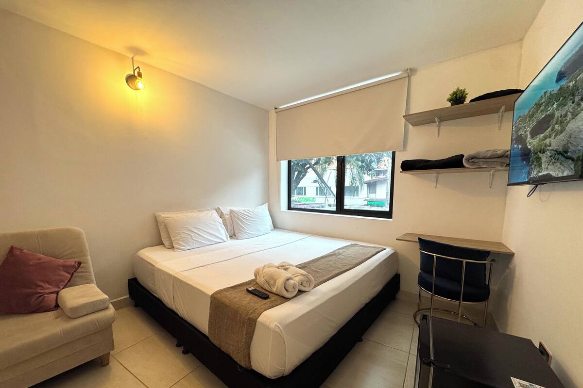 Une chambre avec un lit, un canapé, une télévision et une fenêtre donnant sur des immeubles.