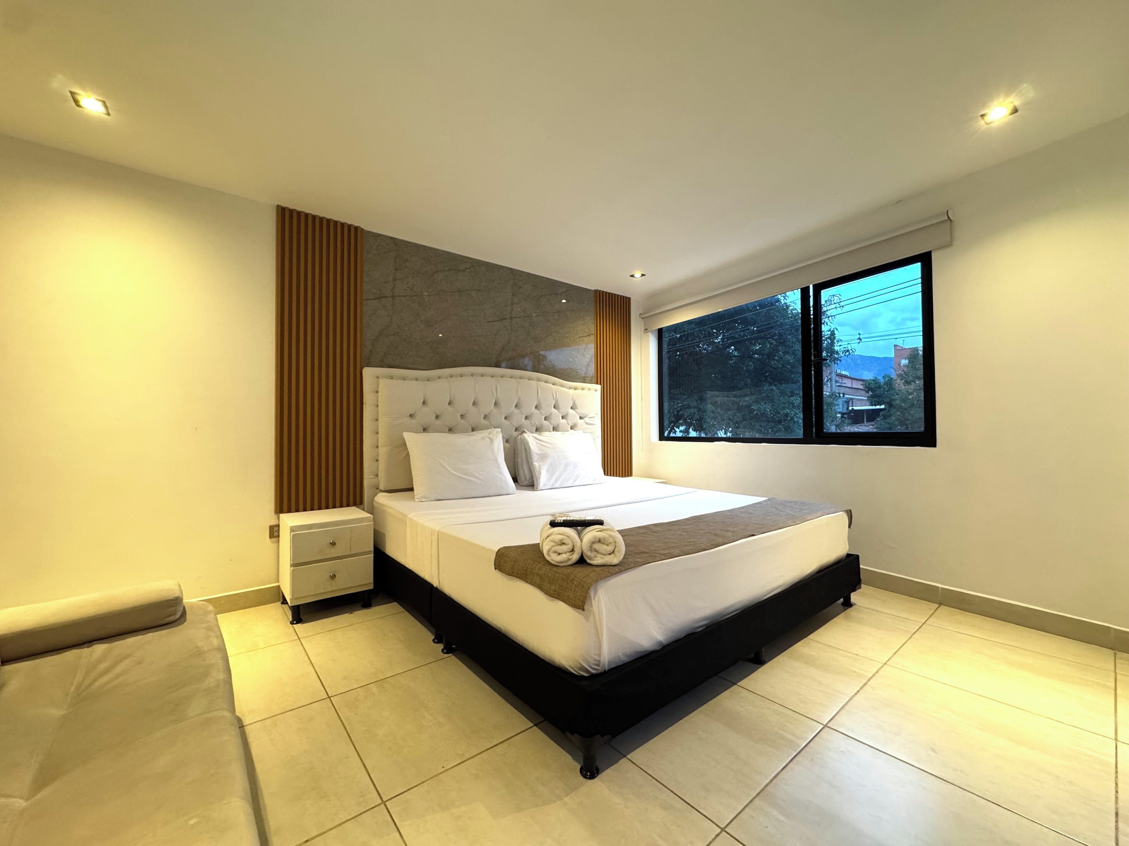 A modern bedroom with a large bed, a nightstand, a window with a view, and a built-in headboard.