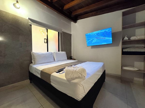 A hotel room with a bed, a television mounted on the wall, and a shelf with folded towels. - URBANITY LIVING (Medellín)