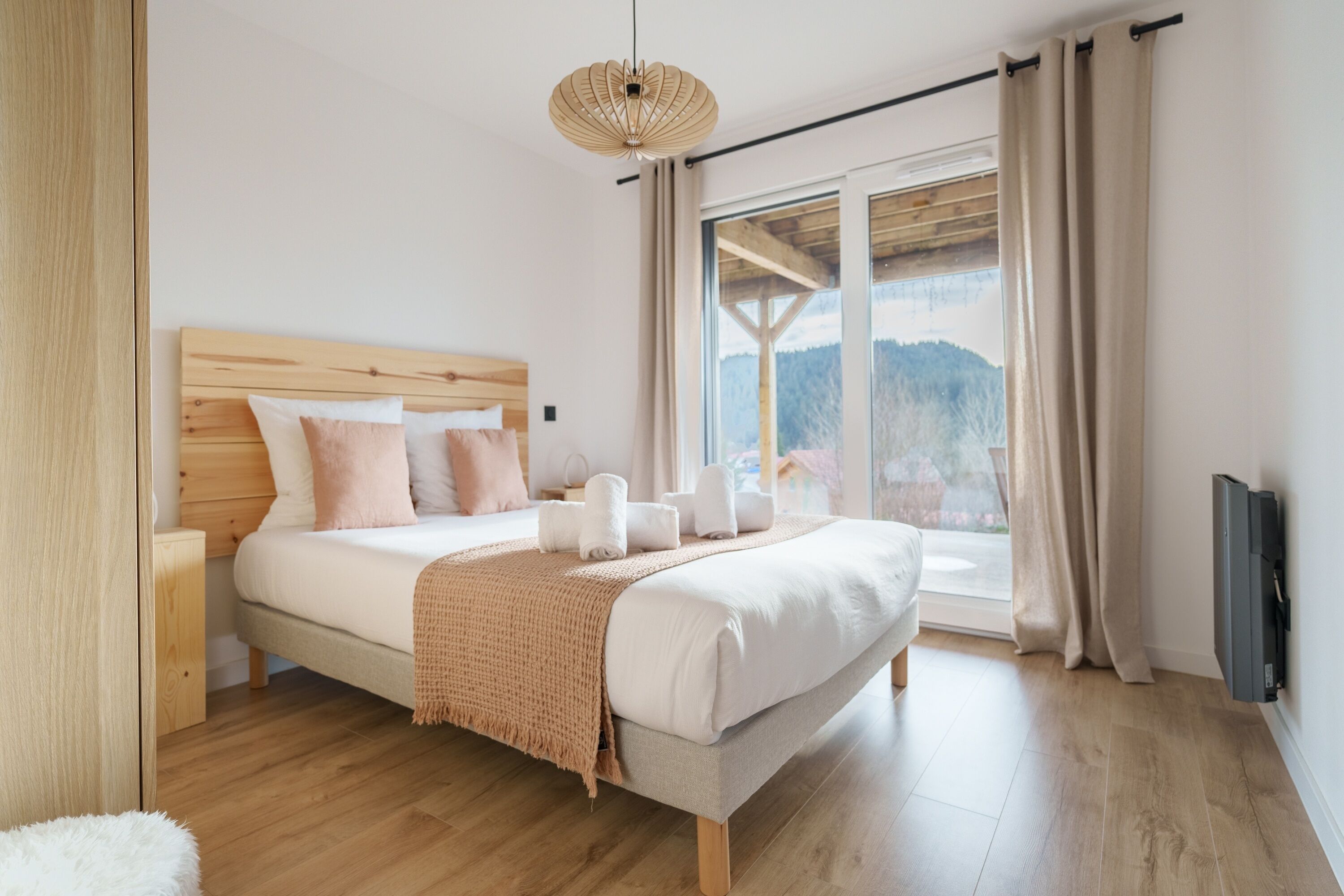 A bedroom with a wooden headboard, a large window with a view, a bedside table, and a TV mounted on the wall.