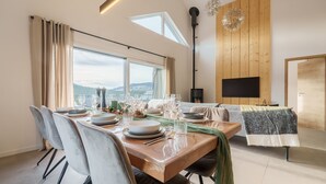 A dining area with a wooden table set for six, large windows, and a flat-screen TV mounted on a wooden wall. - L’Alma - Magnifique maison avec jacuzzi (Gérardmer)