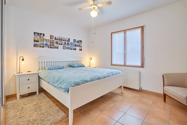 A bedroom with a bed, bedside tables, a ceiling fan, a window, and a wall with photos.
