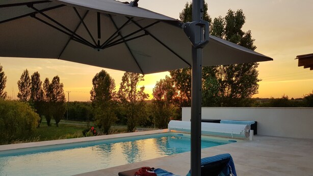A poolside area with lounge chairs, an umbrella, and a view of trees and a sunset.