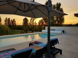A poolside area with lounge chairs, an umbrella, and a view of trees and a sunset.