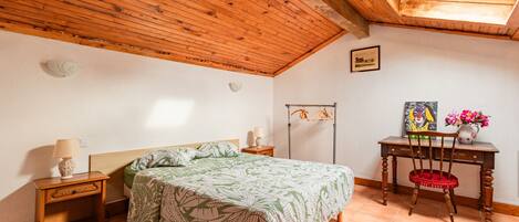 A bedroom with a wooden ceiling, a bed with a green leaf-patterned bedspread, a nightstand with a lamp, a wooden chair, and a desk with a vase of flowers.