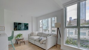 Una sala de estar moderna con un sofá blanco, un sillón verde, una mesita con una planta y un amplio ventanal con vistas a la ciudad.