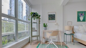 Un dormitorio moderno con un ventanal grande, un escritorio, una silla, una cama y una mesita de noche con una lámpara.