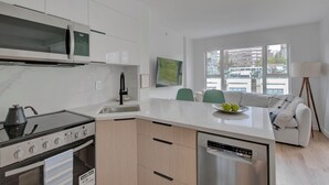 Una cocina moderna con estufa, microondas y lavavajillas. Hay una zona de comedor con sofá y ventana con vistas a la ciudad.