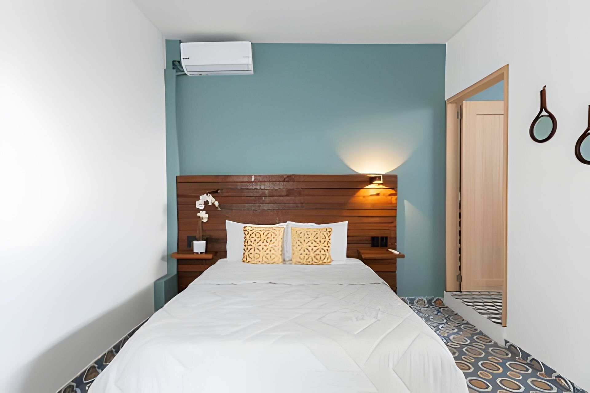 A bedroom with a wooden headboard, a white bedspread, patterned pillows, a bedside lamp, and an air conditioning unit on the wall.