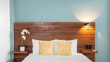 A bedroom with a wooden headboard, two patterned pillows, a white bedspread, a nightstand with a potted plant, and a wall-mounted light.