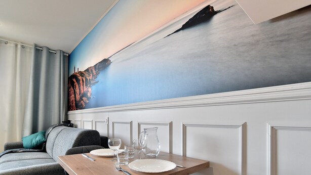 A dining area with a table set for two, a mural on the wall, and a sofa with a cushion. - Paulette 14 - A Sables d'Or les pins, proche plage (Fréhel)
