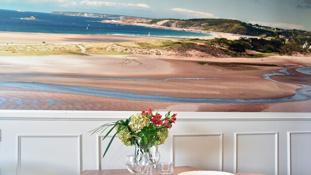Una mesa de comedor con vista al mar, un florero con flores y un paisaje costero de fondo.