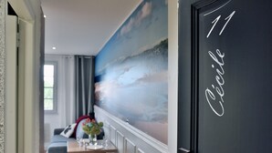Una habitación con un amplio mural de una escena de playa, una zona de comedor con una mesa dispuesta para dos personas, y una puerta con el número 11 y la palabra 'Céline' escrita en ella.