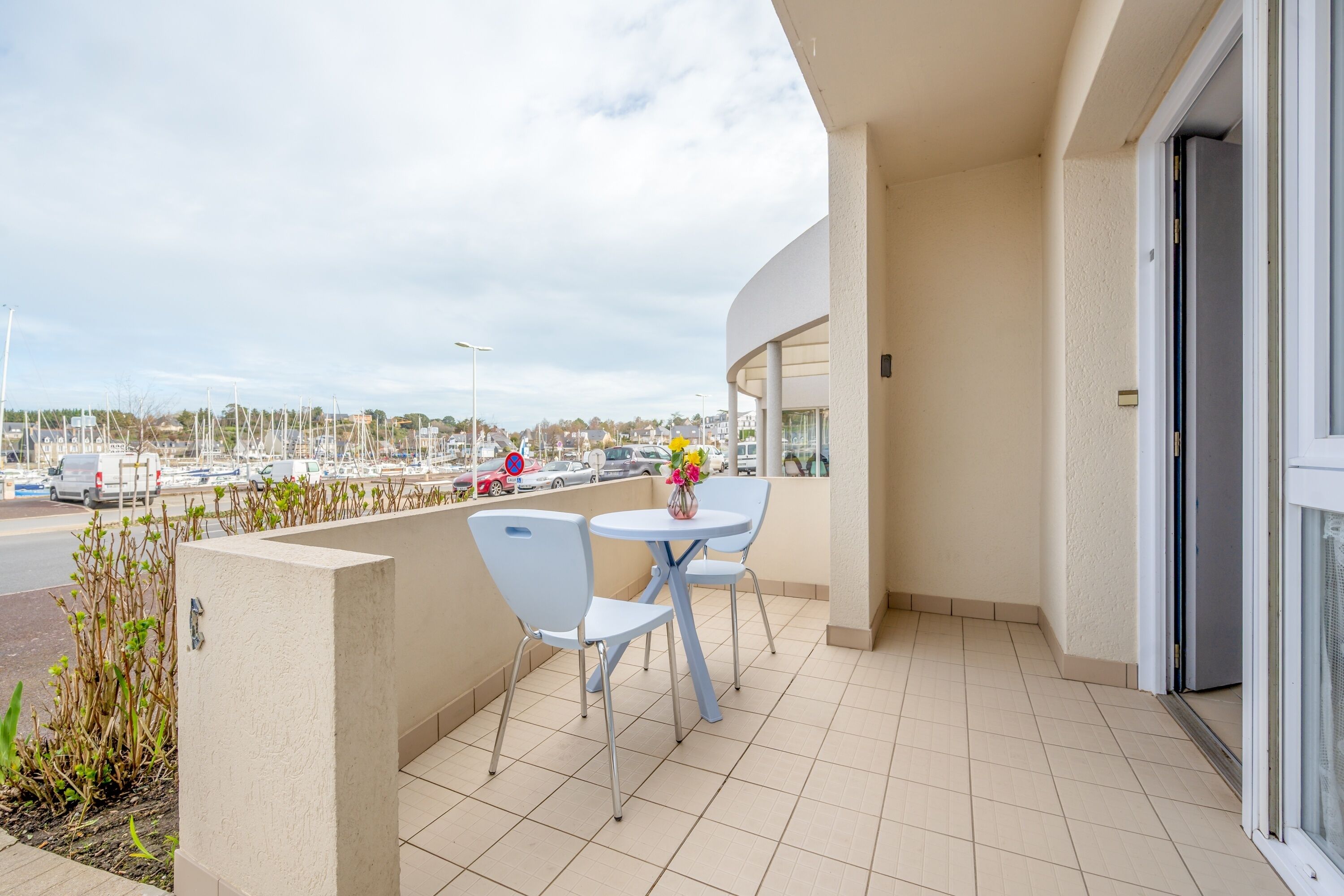 A balcony with two white chairs and a small table, overlooking a marina with boats and a road with cars.