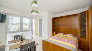Un dormitorio con cama de madera, una mesita con una botella y vasos, y vistas a barcos a través de la ventana.