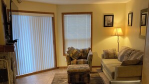 A living room with a sofa, ottoman, and a coffee table. There is a window with blinds, a lamp, and a television. - John's Aspens Ski In/Out (Whistler)