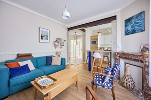 A modern living room with a blue sofa, wooden coffee table, and patterned armchair. A kitchen is visible through an open doorway.