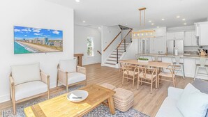 A spacious living area with a wooden coffee table, two white sofas, and a dining table with chairs. A staircase leads to a kitchen with white cabinets and a hanging light fixture.