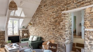 A cozy room with a stone wall, a green sofa, a dining table set with plates and glasses, and a television. - Rêve Emeraude - 100m de la Plage (Cancale)