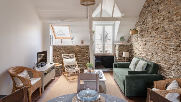 A cozy living room with a stone wall, a green sofa, wicker chairs, and a dining area with a table and chairs. - Rêve Emeraude - 100m de la Plage (Cancale)