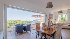 Un comedor con mesa y sillas, un ventanal con vistas a la vegetación y un balcón con sombrilla.